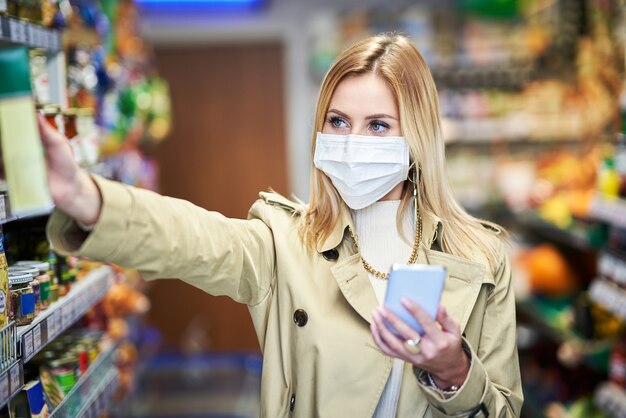 Взрослая женщина в медицинской маске с помощью смартфона и покупки продуктов