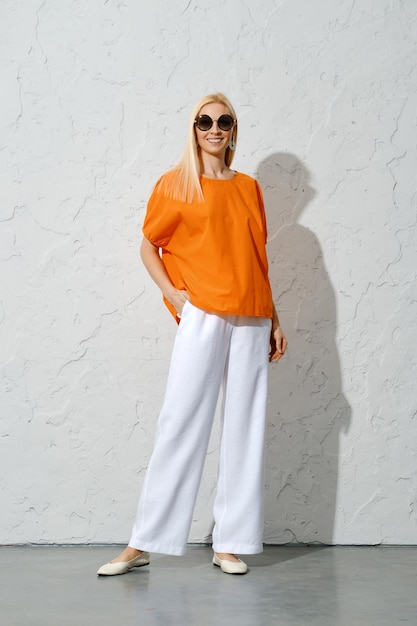 An adult woman posing in a stylish orange blouse and white trousers