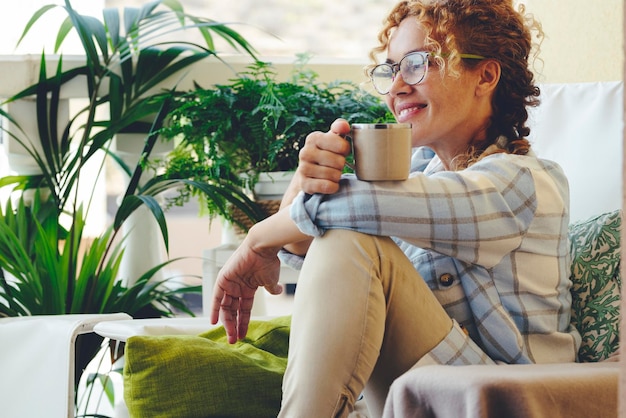 大人の女性がラウンジチェアに座って微笑んで、コーヒーやお茶を飲みながらリラックスした時間を過ごします。眼鏡をかけた女性のための幸せなリラックスしたライフスタイル