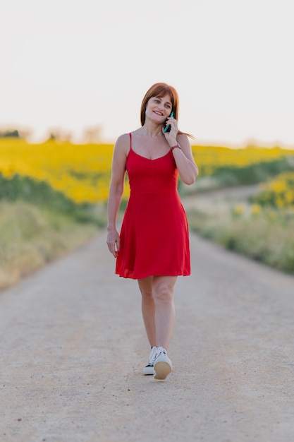 Adult woman wearing a red dress walking in a sunflower field and talking on her mobile phone.