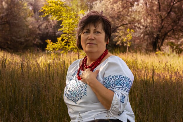 An adult woman in a white embroidered shirt sits in a field against a background of trees. A woman in an old Ukrainian national dress.