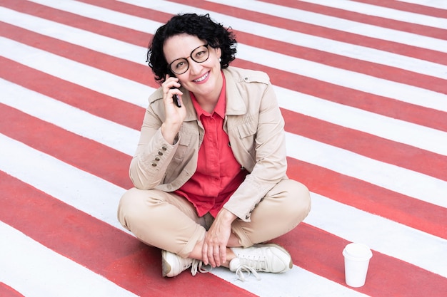 an adult woman with glasses sits on the sidewalk talking on the phone with a smile on her face