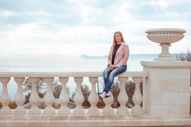 An adult woman with long blond hair sits on a parapet against the backdrop of the Crimean bridge