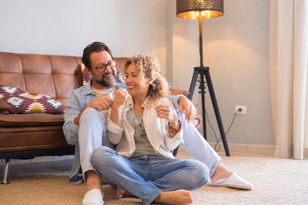 Adult young couple man and woman sitting on the floor at home enjoying in leisure indoor activity together - love and relationship middle age people having relax time in living room