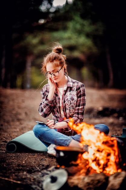 Adult young red hair woman wearing glasses and headphones enjoy listening to music while looking at the phone in forest near the fire