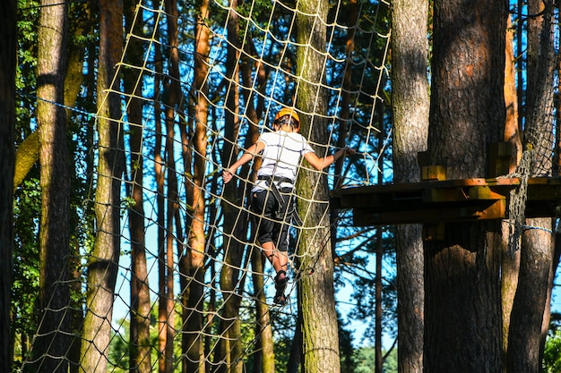 ハイワイヤーパークを登るアドベンチャー。閉じる。ヘルメットをかぶった少年は、冒険公園で楽しんで遊んだり、ロープを持ったり、木製の階段を登ったりしています。アクティブなライフスタイルのコンセプト。夏のレジャー活動