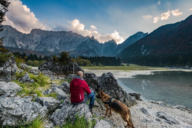 Авантюрист с собакой сидит у скалы и смотрит на альпийское озеро
