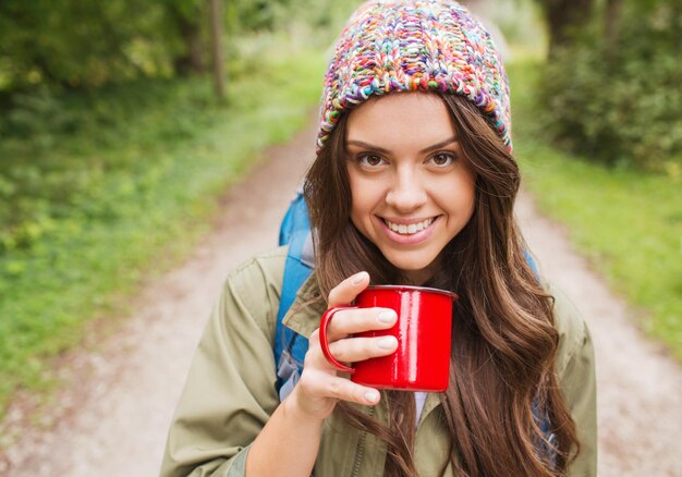 Приключения, путешествия, туризм, походы и концепция людей - улыбающаяся молодая женщина с чашкой и рюкзаком в лесу