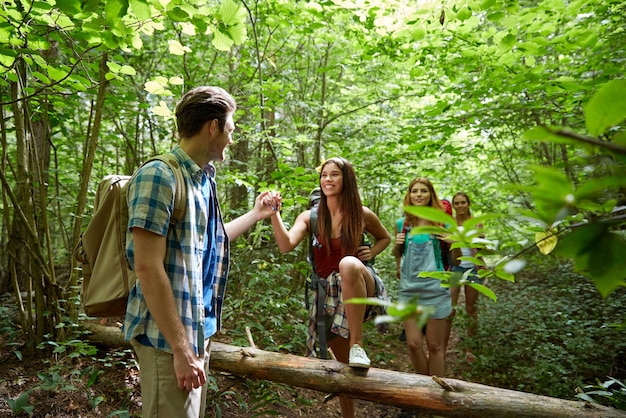 adventure, travel, tourism, hike and people concept - group of smiling friends walking with backpacks and climbing over fallen tree trunk in woods