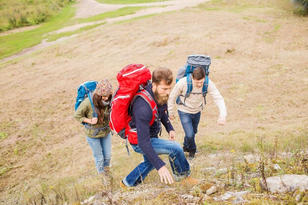приключения, путешествия, туризм, походы и концепция людей - группа улыбающихся друзей с рюкзаками, взбирающихся на холм