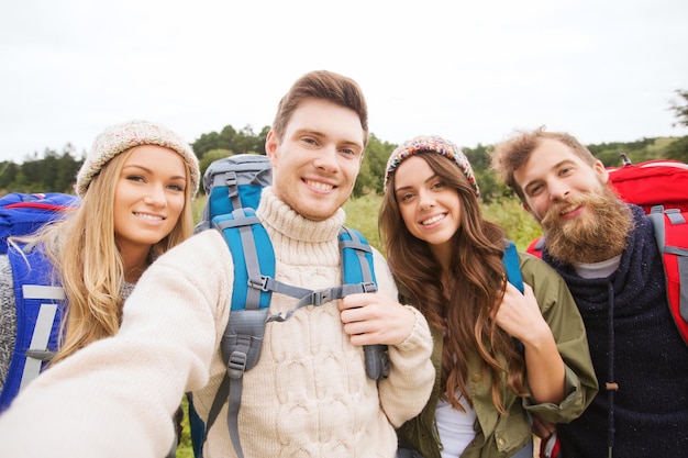 adventure, travel, tourism, hike and people concept - group of smiling friends with backpacks making selfie outdoors