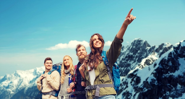 adventure, travel, tourism, hike and people concept - group of smiling friends with backpacks pointing finger over alpine mountains background