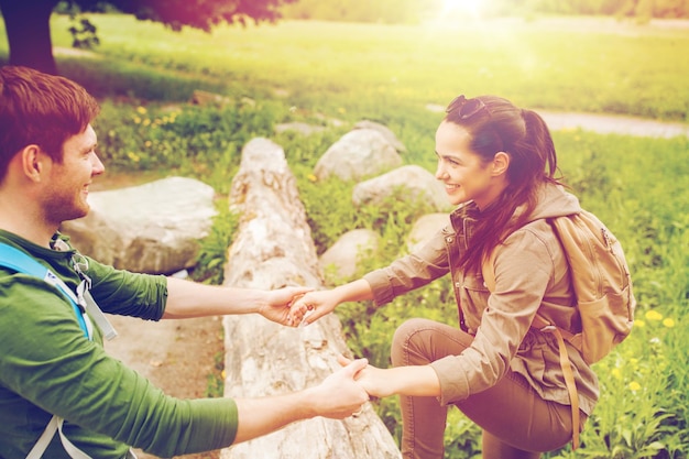 adventure, travel, tourism, hike and people concept - smiling couple with backpacks walking and climbing over fallen tree trunk in woods