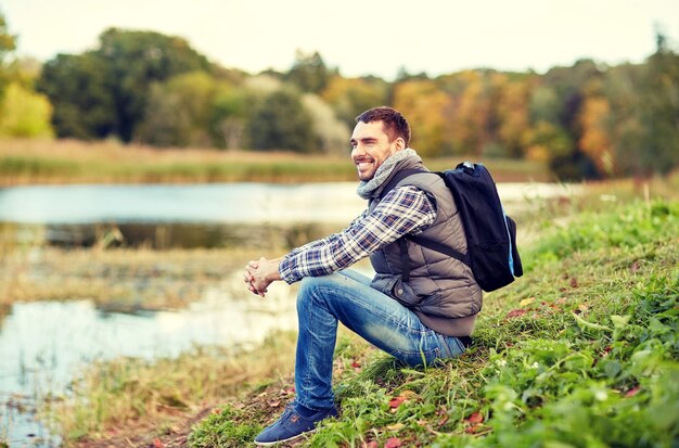 adventure, travel, tourism, hike and people concept - smiling man with backpack resting on river bank