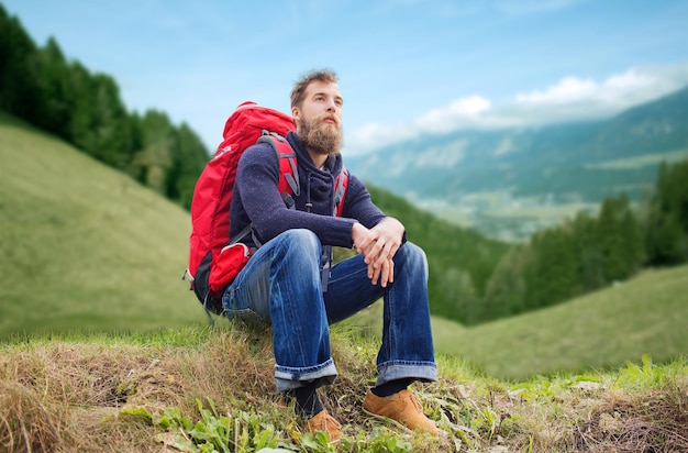 adventure, travel, tourism, hike and people concept - smiling man with red backpack sitting on ground over alpine hills background