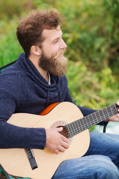 모험, 여행, 관광, 음악 및 사람들 개념 - 캠핑에서 기타를 치는 웃는 남자