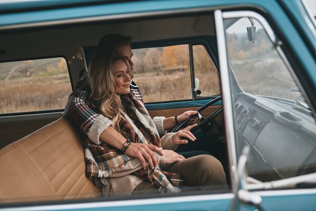 Adventurous couple. Beautiful young couple embracing and smiling while sitting in blue retro style mini van