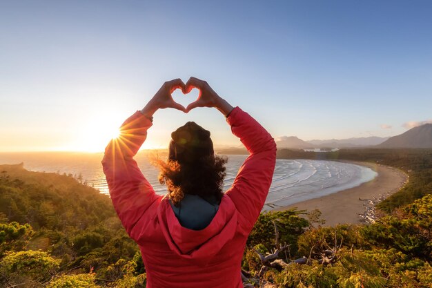 Adventurous woman hiker making heart shape with hands at sandy beach on the west coast