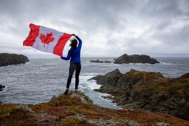 Adventurous woman holding a Canadian Flag on a Rocky Atlantic Ocean Coast