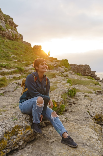 冒険好きな女性は日没時に崖に座っています。山を旅するブルネットの女性の垂直方向のビュー。