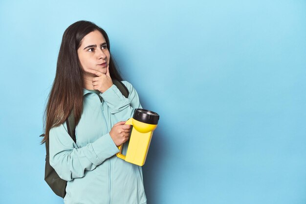 adventurous woman with backpack on blue looking sideways with doubtful and skeptical expression