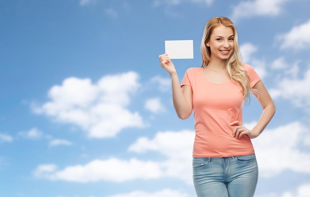 advertentie, uitnodiging, bericht en mensenconcept - lachende jonge vrouw of tienermeisje met lege witte papieren kaart over blauwe lucht en wolken achtergrond