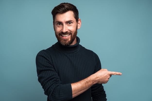 여기에서 광고하십시오. 턱수염을 기른 긍정적인 남자가 광고를 위해 빈 공간에 주의를 기울이고 이빨 미소로 카메라를 바라보며 손가락을 가리키고 있습니다. 파란색 배경에 고립 된 실내 스튜디오 촬영