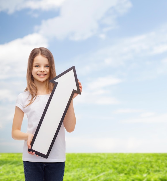 реклама, направление, летние каникулы и концепция детства - улыбающаяся маленькая девочка с белой стрелкой, указывающей вверх на естественном фоне