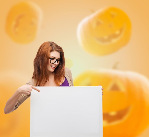 advertising, education, holidays and people concept - smiling teenage girl in glasses pointing finger to blank white board over halloween pumpkins background