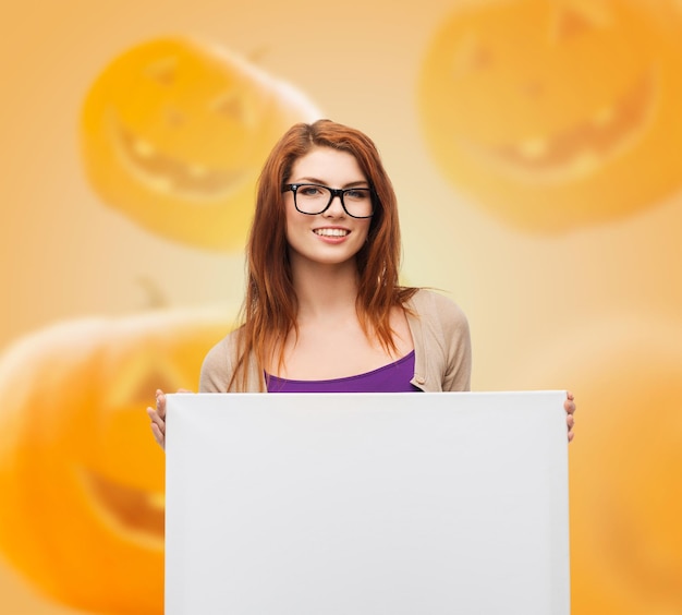 advertising, education, holidays and people concept - smiling teenage girl in glasses with blank white board over halloween pumpkins background