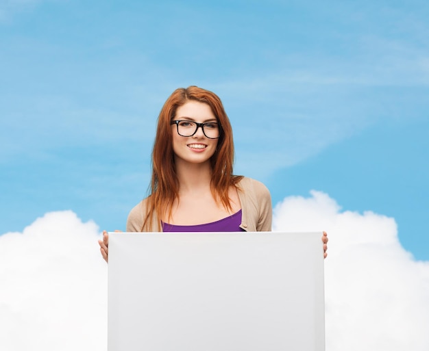 광고, 교육 및 사람 개념 - 푸른 하늘과 구름 배경 위에 빈 흰색 보드가 있는 안경을 쓰고 웃고 있는 10대 소녀