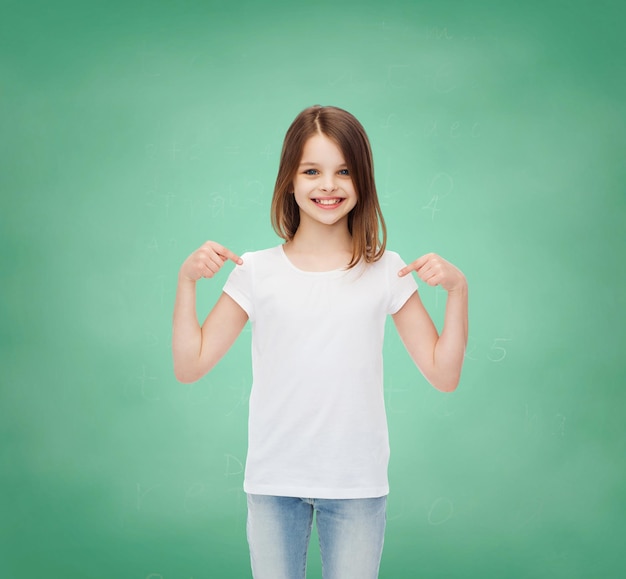 광고, 제스처, 교육, 어린 시절 및 사람들 - 흰색 티셔츠를 입은 웃는 소녀가 녹색 보드 배경 위에 자신을 손가락으로 가리키고 있습니다.