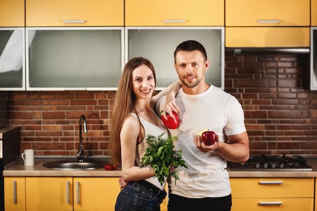 Advertising healthy food, cute young people posing in the kitchen studio. Man and woman holding vegetables and fruits in their hands, smiling and looking at the camera