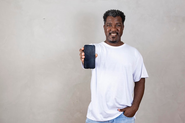 모바일 앱에 광고 회색 스튜디오 배경 위에 포즈를 취한 흰색 빈 스마트폰 화면을 가리키며 카메라를 바라보며 웃고 있는 흥분한 잘생긴 아프리카 남자