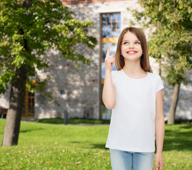 advertising, summer vacation, gesture, childhood and people - smiling little girl in white t-shirt pointing finger up over campus background