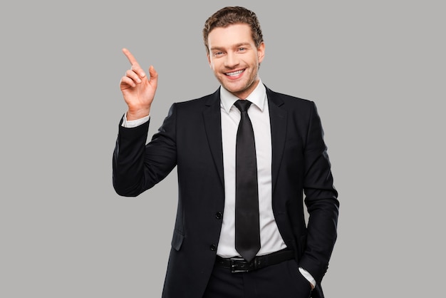 Advertising your product. Confident young man in formalwear looking at camera and smiling while pointing away and standing against grey background