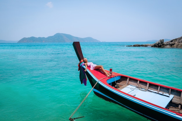 Aerial drone shot of a young blonde woman in a pink dress and sunglasses in the front of a wooden longtail  thai boat. Crystal clear water and corals at a tropical island and amazing beach.