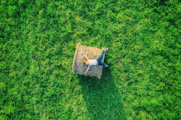 Aerial drone view of happy smiling girl with jeans resting on hay bale and green grass meadow.