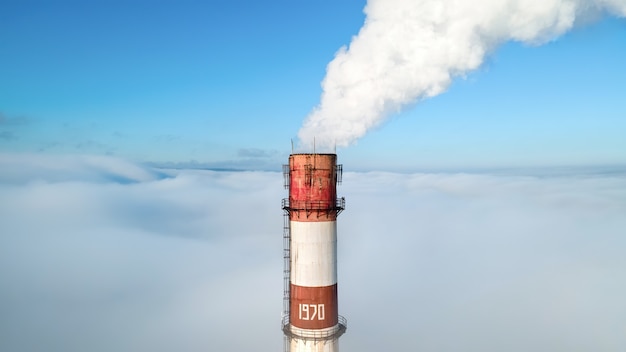 Aerial drone view of thermal station's tube visible above the clouds with smoke coming out. Blue and clear sky