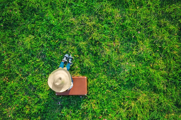 Aerial drone view of tired girl with straw hat and jeans sitting and resting on brown vintage suitcase on green grass meadow. Travel concept.