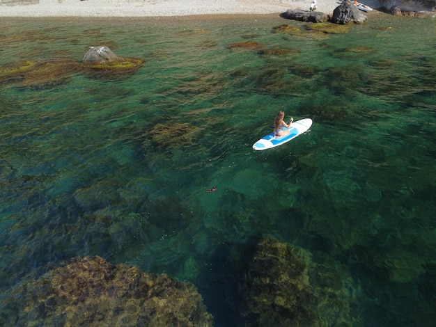 Aerial drone view on young attractive brunette woman with long hair in blue swimsuit swimming on sup around volcanic rocks like in Iceland Summer holiday vacation and travel concept