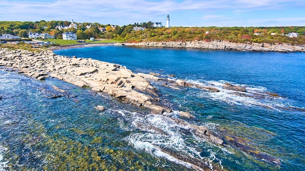 Aerial over ocean and coast with shallow water in Maine and lighthouse with homes in background