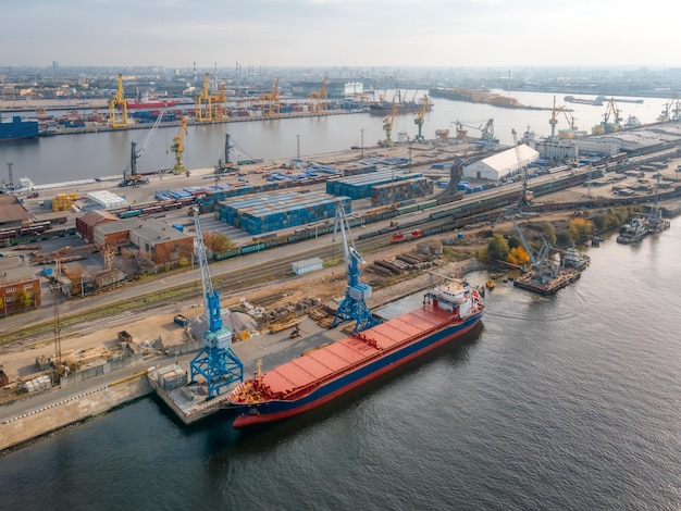 Aerial photo of port with boats, cranes and buildings. Port cranes load various cargoes into the hold of the red ship. Goods delivery by sea. On the background railway wagons and containers.