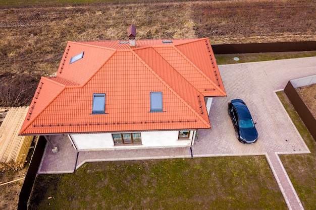 Aerial top view of house metal shingle roof with attic windows and black car on paved yard.