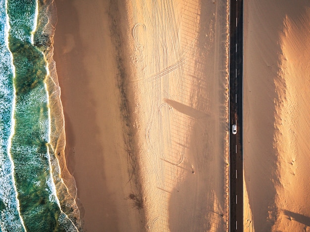 Aerial vertical view of yellow tropical sandy beach with black long way road and car traveling - ocean blue waves and shore - sunset time with long beautiful shadow - concept of summer vacation