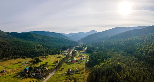 Photo aerial view of amazing scenery with small village houses between foggy dark mountain peaks covered with forest pine trees at autumn sunrise beautiful wild woodland with shining rays of light at dawn