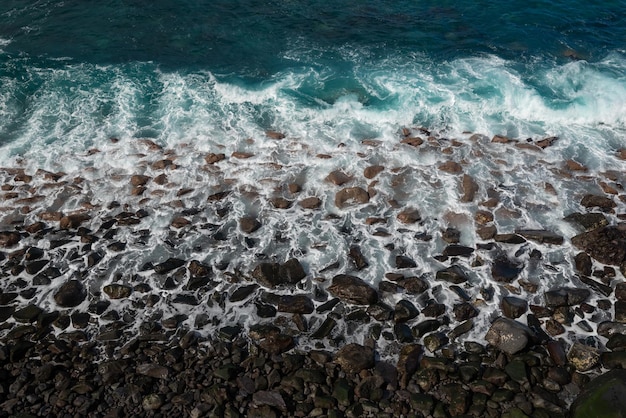 Aerial view of black beach with wave splashing over volcanic rocks Bright turquoise blue ocean Coastline photography Top view of ocean shore White pattern of sea foam on stones Marine photo