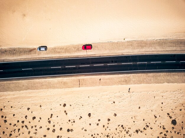 両側に砂と砂漠がある黒いまっすぐなアスファルト道路の航空写真-側面に2台の車が駐車されています-エキゾチックな目的地のための旅行と放浪癖の概念