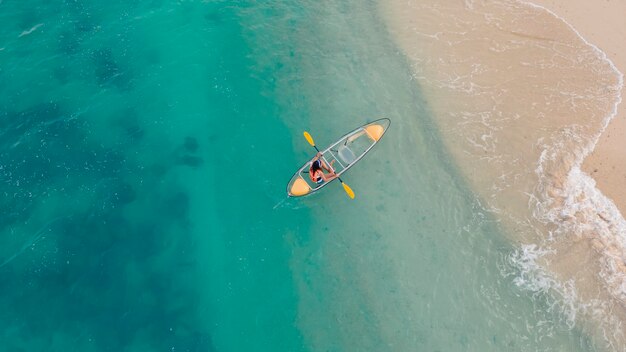 Aerial view of blue sea with floating transparent kayak is going into sand beach, person with life jacket on boat