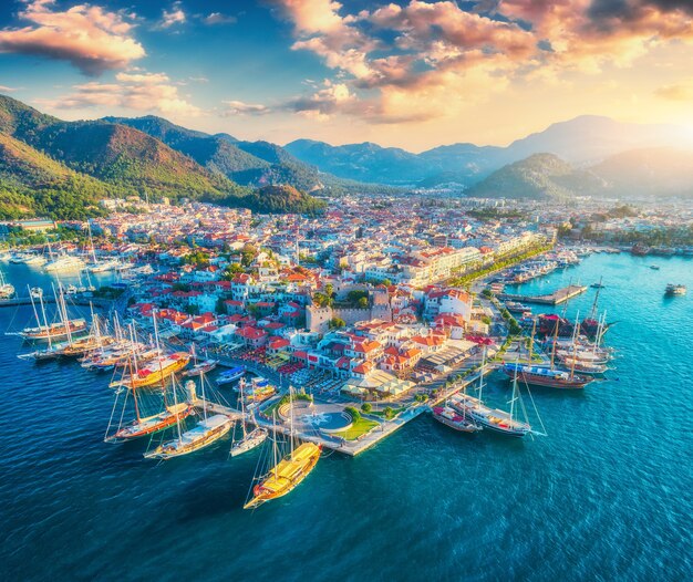 Aerial view of boats and yachts and beautiful architecture at sunset in Marmaris, Turkey. Landscape with boats in blue sea, houses, city, mountains, colorful sky with clouds. Top view of harbor